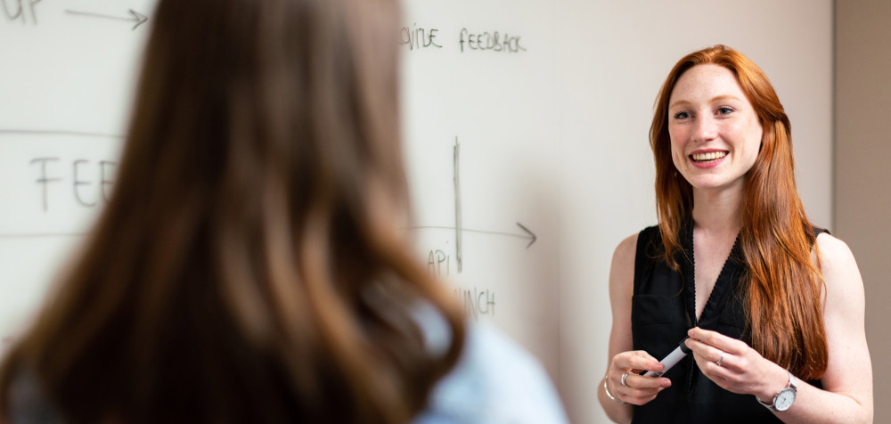 Eine junge Frau steht vor einem Whiteboard und erklärt einer anderen etwas.