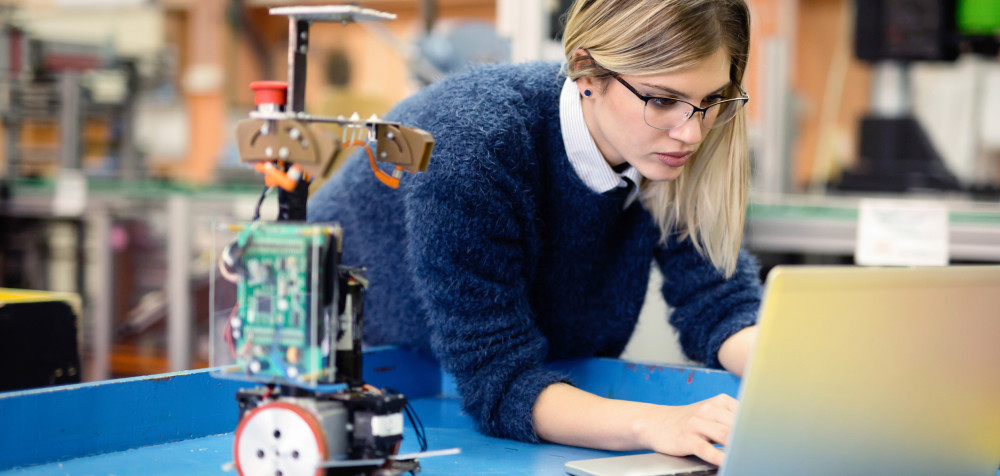 Ein junge Frau arbeitet an einem Laptop. Daneben steht ein kleiner Roboter.