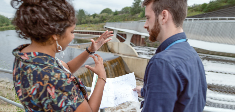 Eine Frau und ein Mann stehen an einem Wehr mit einer Brücke an mit Plänen in der Hand.