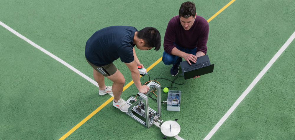 Zwei junge Studierende befinden sich auf einem Tennisplatz. Sie stellen eine Ballmaschine ein.