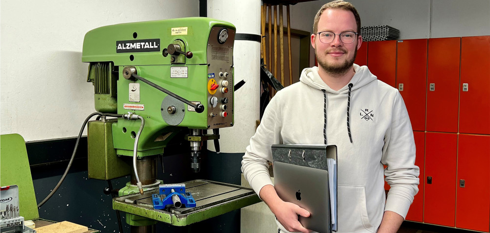 Junger Mann steht vor einer Industriemaschine mit Ordner und Macbook in der Hand