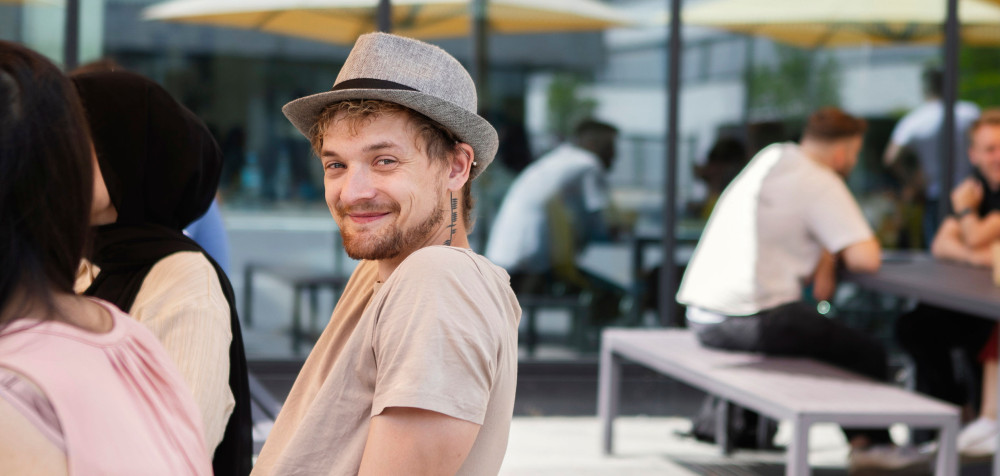 Ein junger Mann sitzt gemeinsam mit Studierenden auf einer Bank vor der Mensa. 